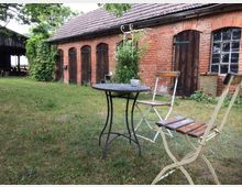 Garten mit einem kleinen runden Tisch aus Metall und zwei klappbaren Holzstühlen, vor einem alten Backsteingebäude mit dunklen Holztüren und Fenstern. Im Hintergrund steht eine überdachte Holzscheune, und der Garten ist von Gras und rankenden Pflanzen bewachsen.