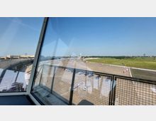 Blick aus einem verglasten Raum auf das ehemalige Flughafengelände Tempelhof in Berlin, mit einer großen betonierten Fläche, umliegenden Grünflächen und dem markanten Flughafengebäude im Hintergrund. Ein weißer Kontrollturm ist ebenfalls sichtbar, unter einem klaren blauen Himmel.