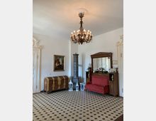 Historischer Innenraum mit einer Kronleuchter-Deckenlampe, einer roten Polsterbank, einem verzierten Kachelofen und einer reich dekorierten Holztruhe. Der Raum hat schwarz-weiß gemusterten Boden und ist mit weiteren antiken Möbelstücken und Dekorationen geschmückt.