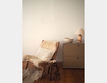 Cozy indoor corner with a wicker lounge chair draped in cream sheepskin and a blanket on a wooden floor, beside a small wooden stool. A low metal cabinet against a white textured wall holds a stone bowl and a table lamp.