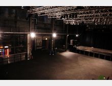 Innenraum eines Veranstaltungsortes mit industriellem Design, hohen Metallkonstruktionen und einem leeren, dunklen Bühnenbereich. Deckenstruktur mit zahlreichen montierten Scheinwerfern, karger Boden, und links eine abgetrennte Bereich mit einem beleuchteten Kühlschrank.