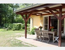 Eine überdachte Terrasse mit Holzkonstruktion an einem gelben Haus, ausgestattet mit einem Holztisch, mehreren Holzstühlen und Blumenvase. Im Hintergrund ein Garten mit Rasen, Hecken, einem Gewächshaus und einer Sonnenliege.
