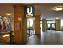 U-Bahnhof mit gelb gekachelten Wänden, einer beleuchteten Beschilderung mit der Aufschrift „U6“ und einem Treppenabgang. Im Hintergrund sind Glastüren und Fenster zu sehen, die Tageslicht in den Raum lassen.