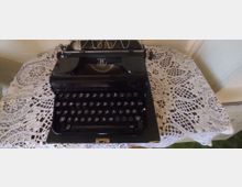 Schwarze Schreibmaschine im Retro-Stil, platziert auf einem mit einem weißen, gehäkelten Spitzentuch bedeckten Tisch. Im Hintergrund sind helle Wände und ein Teil eines Heizkörpers sichtbar.