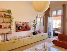 Wohnzimmer mit hellen Wänden, einer gelben Kommode, darauf Bücher und Dekorationen, sowie an der Wand ein Kunstbild und ein rundes Spiegel. Rechts führt eine geöffnete Glastür auf einen kleinen Balkon mit Sitzmöbeln. Ein großen Papierlampenschirm hängt von der Decke, und ein Sessel sowie ein Couchtisch mit Marmoroptik stehen im Raum.