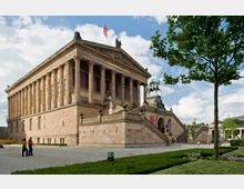 Neoklassizistisches Museumsgebäude mit Säulenfassade, einer breiten Treppe mit Skulpturen und einem Reiterstandbild auf einem Podest. Im Vordergrund befinden sich ein gepflasterter Platz und gepflegte Grünanlagen mit Bäumen.