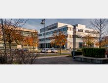 Moderne Bürogebäude mit großen Fenstern, umgeben von herbstlich gefärbten Bäumen und geparkten Autos auf einer gepflasterten Straße. Im Vordergrund Betonbänke und Gehwege, die zu einer urbanen Umgebung passen.