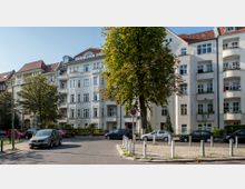 Straßenszene mit mehrstöckigen, weiß verputzten Wohnhäusern im klassischen Altbaustil, teilweise mit Erkern und Balkonen. Im Vordergrund sind geparkte Autos, ein großer Baum und Begrenzungspfosten auf einem Platz zu sehen.