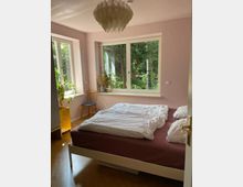 Schlafzimmer mit Holzfußboden, einem Doppelbett mit gemusterter Bettwäsche und großen Fenstern, die einen Garten mit Bäumen zeigen. Ein dekorativer Kronleuchter hängt von der Decke, und Pflanzen sowie Möbelstücke sind dezent im Raum verteilt.