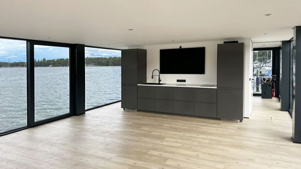 Modernes Interieur eines schwimmenden Hauses oder Gebäudes mit großen bodentiefen Fenstern, die auf eine Wasserlandschaft blicken. Im Raum befindet sich eine dunkelgraue Küchenzeile mit integriertem Waschbecken und flachem Bildschirm; der Boden besteht aus hellem Holz.