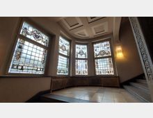 Treppenhausbereich in einem historischen Gebäude mit großen Buntglasfenstern, die kunstvolle Verzierungen und Wappen zeigen. Der Raum hat einen gefliesten Boden, ein verziertes Geländer und eine Decke mit Kassettendesign.