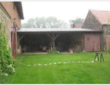 Ein ländlicher Hof mit einem offenen Schuppen mit Wellblechdach, umgeben von Ziegel- und Feldsteinmauern. Im Vordergrund ist eine gepflegte Rasenfläche mit weißem Absperrband sichtbar, während im Hintergrund Bäume im Regen zu erkennen sind.