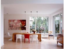 Modernes Esszimmer mit hellem Holzboden und einem langen Holztisch, umgeben von weißen und einem roten Stuhl. Große Fensterfronten bieten Blick auf einen grünen Garten, und eine farbenfrohe Kunstleinwand hängt an der Wand links.