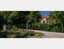 Eine Statue eines stehenden Mannes auf einem Steinsockel in einem Park mit blühenden Blumenbeeten und einem geschwungenen Kiesweg. Im Hintergrund befindet sich ein Gebäude mit heller Fassade und einem roten Ziegeldach, umgeben von dichter grüner Vegetation.