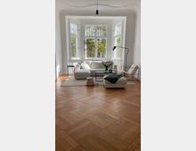 Wohnzimmer mit großem Erkerfenster aus mehreren weißen Sprossenfenstern, hellem Holzparkett und modernen Möbeln in neutralen Farben, darunter ein Sofa, ein Sessel, ein Couchtisch und eine Stehlampe. Durch die Fenster ist grüne Vegetation zu sehen.