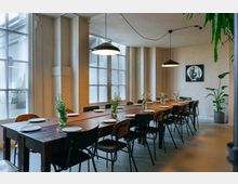 Ein geschmackvoll eingerichteter Speiseraum mit einem langen Holztisch, umgeben von schwarzen und braunen Stühlen. Auf dem Tisch stehen Teller, Besteck und Vasen mit Blumen; im Hintergrund große Fenster und dezente Wanddekoration mit einem Schwarz-Weiß-Bild.