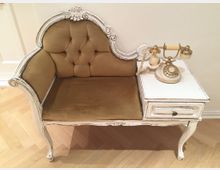 Weiß lackiertes, antikes Möbelstück mit gepolstertem Sitzbereich in beigefarbenem Stoff und detailliert geschnitzter Rückenlehne, kombiniert mit einer kleinen Ablagefläche, auf der ein cremefarbenes, retro-inspiriertes Telefon steht; steht auf einem Holzboden.