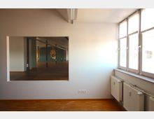 Ein heller Raum mit Holzfußboden, großen Fenstern auf der rechten Seite und einer weiß gestrichenen Wand. In der Wand befindet sich ein rechteckiger Durchbruch, durch den ein größerer Raum mit Säulen, Parkettboden und einer Wand mit einem abstrakten Kunstwerk in Form einer Hand sichtbar ist.