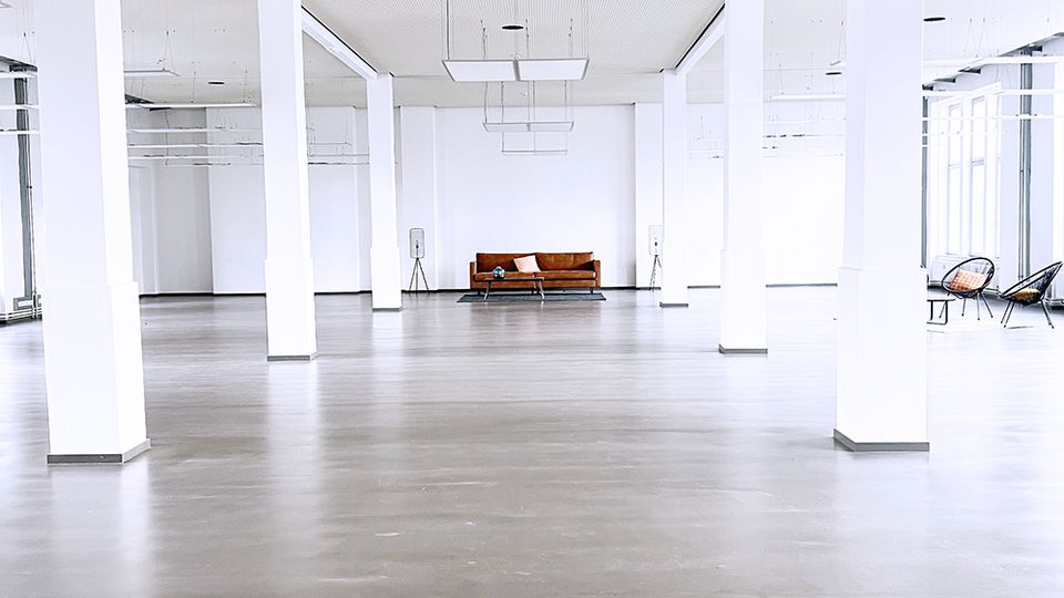 Großer, heller Raum mit weißen Wänden und vielen rechteckigen, weißen Säulen, die den Raum gliedern. Der Boden ist grau und glatt, die Decke perforiert mit rechteckigen Hängelampen; im Hintergrund steht ein einzelnes braunes Sofa.