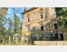 Verlassenes, zweistöckiges Gebäude mit bröckelnder Fassade und teilweise zerstörten Fenstern, umgeben von dichter Vegetation und alten Rohren im Vordergrund. Die Umgebung besteht aus Bäumen und überwuchernden Pflanzen, die das verwitterte Erscheinungsbild unterstreichen.