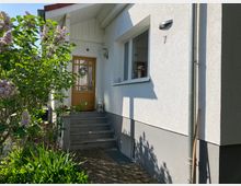 Eingangsbereich eines weißen Einfamilienhauses mit einem kleinen Treppenaufgang und einer hölzernen Eingangstür. Links vom Eingang befindet sich ein blühender Fliederbusch, während der Boden mit Pflastersteinen ausgelegt ist.