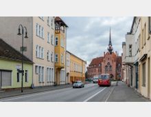 Straßenszene in einer Kleinstadt mit mehrstöckigen Wohngebäuden in Pastellfarben links und rechts der Straße. Im Hintergrund ist ein historisches Gebäude mit rotem Ziegeldach, Spitzgiebel und Turm mit Uhr zu sehen.