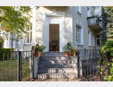 Eingang eines Altbaus mit stufenförmiger Granittreppe, flankiert von Pflanzen in Tontöpfen. Das Gebäude hat cremefarbene Wände, weiße Fensterrahmen und dekorative Stuckelemente.