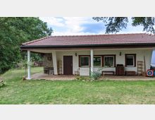 Einstöckiges Gebäude mit einem Ziegeldach und einer überdachten Terrasse, umgeben von einem Garten mit Rasen und Bäumen. Die weiße Fassade hat Holzfensterrahmen, eine zentrale Holztür und dekorative Elemente wie Blumentöpfe und Gartenmöbel.