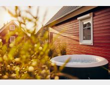 Ein rotes Holzhaus mit einem weißen Fenster, daneben im Vordergrund ein aufblasbarer Whirlpool im Garten. Im Hintergrund sind weitere Häuser sowie Pflanzen in warmem Sonnenlicht zu sehen.