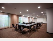 Tagungsraum mit rechteckig angeordneten Tischen und gepolsterten Bürostühlen auf einem braunen Teppichboden. Der Raum ist von Vorhängen umgeben, die bodentiefe Fenster verdecken, und wird durch Deckenleuchten und Tageslicht beleuchtet.