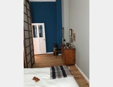 Ein Schlafzimmer mit Holzboden, einer weißen Wand und einer Akzentwand in Blau. Zu sehen sind ein Bett mit einer gestreiften Decke, eine Kommode aus Holz mit dekorativen Gegenständen und gerahmten Bildern sowie eine weiße Tür im Hintergrund.