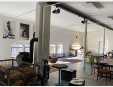 Großzügiger, offener Raum mit industriellem Design, gekennzeichnet durch weiße Backsteinwände, freiliegende Metallstreben und große Rundbogenfenster. Der Raum ist mit unterschiedlichen Möbeln wie Tischen, Stühlen und dekorativen Elementen wie Teppichen, Fotografien sowie einem Ofen ausgestattet.