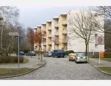 Mehrstöckiger Wohnblock mit hellen Fassaden und kleinen Balkonen, umgeben von Bäumen ohne Laub. Im Vordergrund gepflasterte Straße mit parkenden Autos und einem Tempo-30-Zonen-Schild.