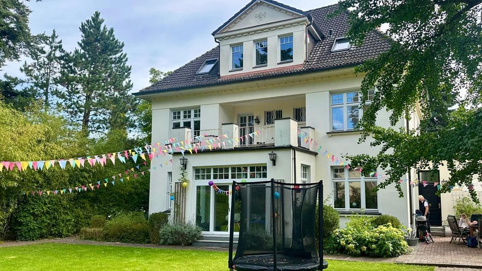 Großes zweistöckiges Einfamilienhaus mit weißer Fassade und Satteldach, umgeben von einem gepflegten Garten mit grünem Rasen, Sträuchern und Bäumen. Im Vordergrund ist ein Trampolin zu sehen, und bunte Wimpelketten schmücken den Garten und die Terrasse.