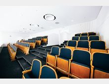 Ein moderner Vortragssaal mit ansteigenden, fest installierten Sitzreihen aus Holz mit dunkelblauer Polsterung. Der Raum hat weiße Wände, einen dunklen Teppichboden und eine helle Decke mit runden Lüftungsöffnungen.