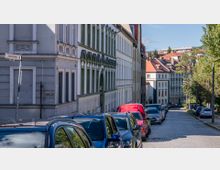 Straßenszene in einer Wohngegend mit historischen Gebäuden, die durch detaillierte Fassaden mit Rundbögen und Ornamenten gekennzeichnet sind. Auf der gepflasterten Straße stehen parkende Autos, und am rechten Rand ist eine Baumreihe sichtbar.