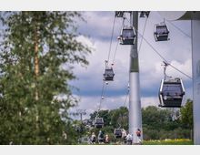 Seilbahn mit mehreren Gondeln, die über eine grüne Landschaft mit Bäumen und Blumen führt; im Vordergrund sind Spaziergänger auf einem Weg zu sehen, während der Himmel von Wolken bedeckt ist.