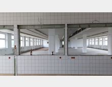 Ehemaliger Fabrik- oder Industriebereich mit leerem Raum, weißen gefliesten Wänden und großen rechteckigen Fensterreihen, die viel Tageslicht hereinlassen; im Vordergrund ist eine Wandöffnung mit Fliesen erkennbar, auf der eine leere Flasche steht.