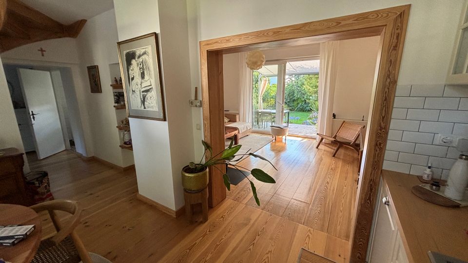 Offener Wohnbereich mit hellem Holzboden und breitem Holztürrahmen, der in einen Sitzbereich mit großen Terrassentüren führt. Rechts ist eine teilweise sichtbare Küche mit weißen Fliesen und Arbeitsplatte, im Hintergrund Blick auf Terrasse und grünen Garten.