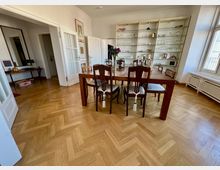 Esszimmer mit Holzboden in Fischgrätmuster, einem langen Holztisch und mehreren gepolsterten Stühlen. Im Hintergrund ein deckenhohes Regal mit Gläsern und Dekorationen, große Fenster auf der rechten Seite lassen Tageslicht herein.