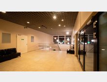 Heller Flurbereich mit Fliesenboden, schwarzen Sitzbänken, einer weißen Tür links und einer großen Glasfassade rechts. Im Hintergrund sind ein Geländer, Fenster mit Blick auf bunte Leuchten und eine Treppe erkennbar.