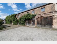 Backsteingebäude mit begrünten Fassaden, alten Holztoren und kleinen Fenstern unter einem schrägen, mit Blech gedeckten Dach, umgeben von einem gepflasterten Hof und freiem Himmel mit Wolken.