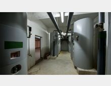 Ein technischer Raum mit mehreren großen, zylindrischen Tanks und Rohrleitungen an der Decke. Die Wände bestehen aus weiß gestrichenem Mauerwerk, und der Boden ist aus Beton.