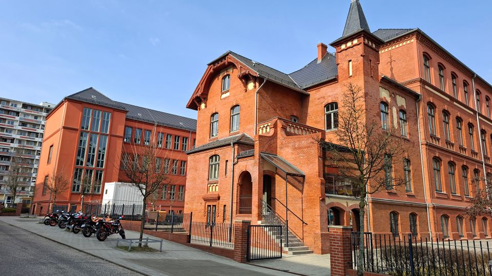 Historisches Backsteingebäude mit mehrstöckiger Fassade, Rundbogenfenstern und einem kleinen Turmaufsatz, daneben ein modernerer Anbau mit großen Fensterflächen. Davor liegen ein gepflasterter Hof mit Zaun, einigen kahlen Bäumen und abgestellten Motorrädern; im Hintergrund ist ein hohes Wohnhaus zu sehen.