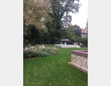 Grüne Gartenlandschaft mit einem gepflegten Rasen, einem kleinen Blumenbeet links im Vordergrund und großen Bäumen im Hintergrund. Im hinteren Bereich befindet sich eine erhöhte Terrasse mit Pool, Sonnenliegen und einer modernen Gartenüberdachung, flankiert von weiteren Pflanzen.