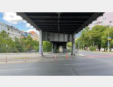 Eine Straßenszene unter einer erhöhten Stahlbrücke mit sichtbaren Querträgern, flankiert von Bäumen und mehrstöckigen Wohnhäusern im Hintergrund. Im Vordergrund ist eine Straße mit Bordsteinkanten und vereinzelten Absperrpylonen zu sehen.