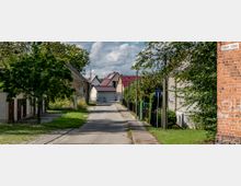 Schmale, gepflasterte Straße in einem Dorf mit Wohnhäusern auf beiden Seiten, teils aus Backstein und mit roten Dächern. Bäume und Sträucher säumen die Straße, rechts im Bild ein Sackgassenschild und eine Mauer aus Ziegelsteinen.