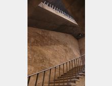 Treppenhaus mit modernem Design, bestehend aus Steinwänden in einem beige-braunen Farbton und Metallgeländern. Sichtbar sind zwei Treppenabschnitte, der obere führt zu einer Zwischendecke mit Geländer.