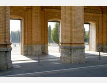 Backsteinarkade mit massiven Säulen und verzierten Bögen; im Hintergrund sind ein gepflasterter Gehweg, Bäume und eine offene Fläche sichtbar. Die Wände bestehen aus roten Ziegeln, teils mit Steinblöcken verstärkt, und die Umgebung wirkt städtisch.