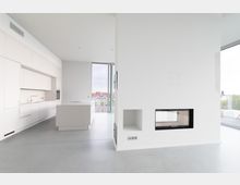 Minimalistisches, modernes Interieur mit einer weißen Küche, bestehend aus Einbauschränken, einem Kochfeld auf einer Insel sowie einer großen Fensterfront mit Blick auf Häuserdächer. Im Vordergrund eine weiße Wand mit einem eingebauten Kamin und Regalfächern.